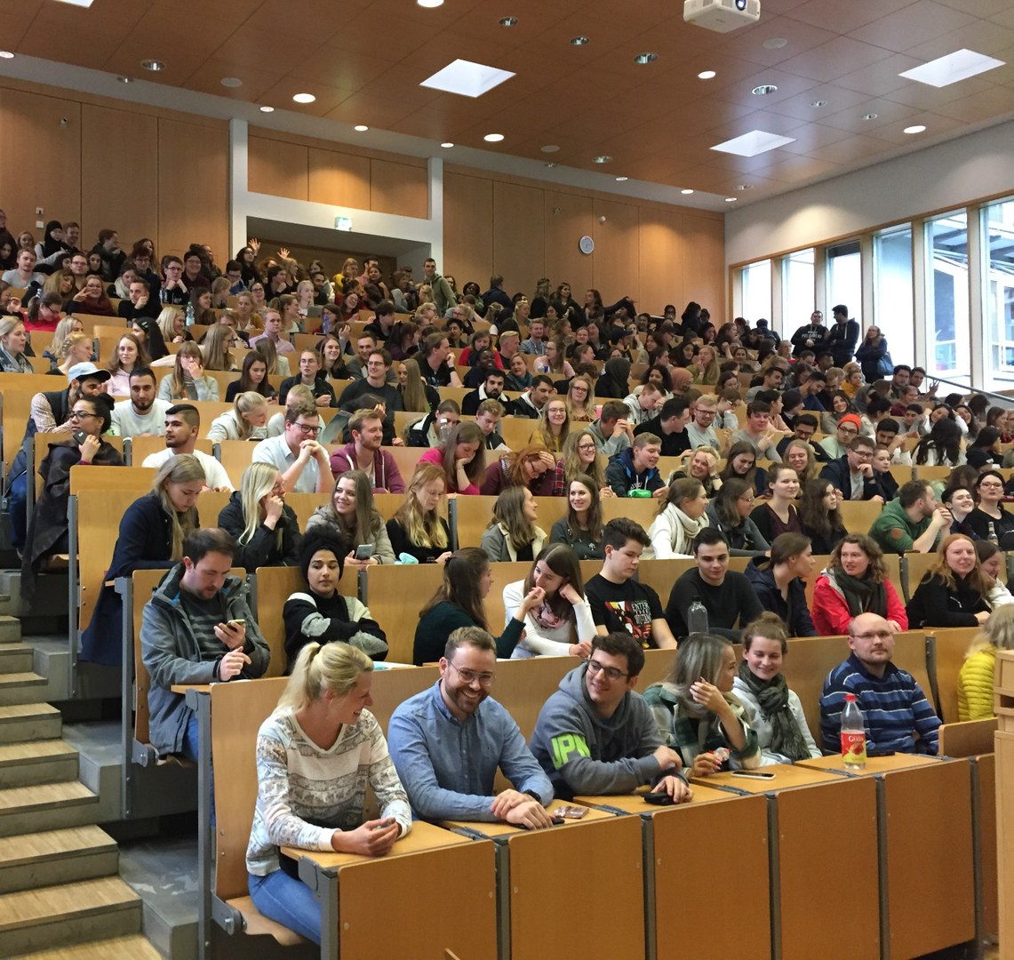 Eine Wissenschaftlerin und ein Wissenschaftler arbeiten hinter einer Glasfassade und mischen Chemikalien mit Großgeräten.
